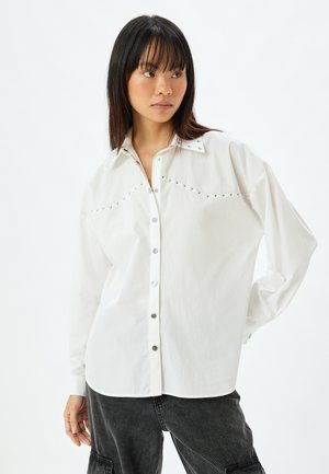 Camicia bianca con bottoni automatici, borchie decorative lungo il collo e maniche lunghe dal fit morbido. Tessuto liscio e leggero.