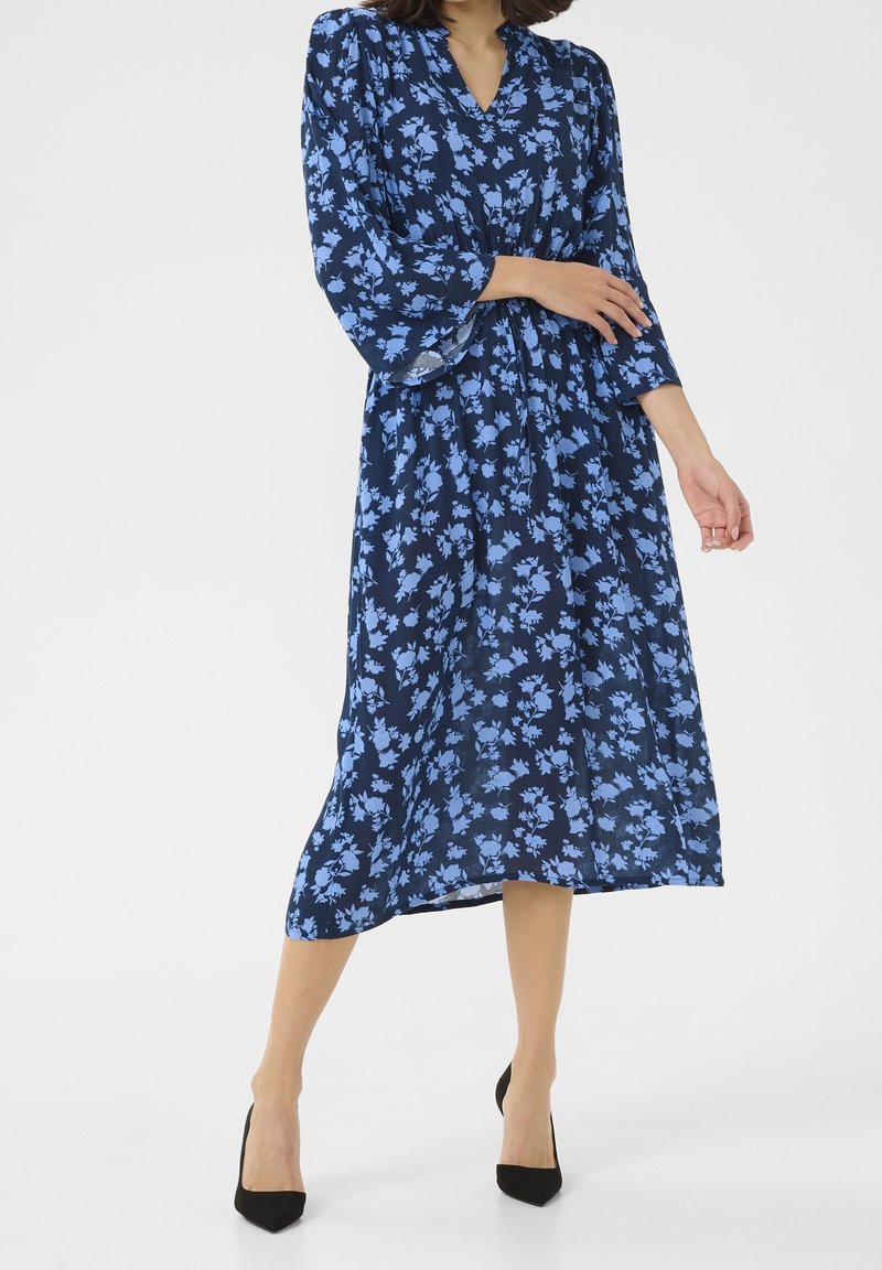 Femme portant une robe midi bleu marine à imprimé floral bleu clair, manches longues, décolleté en V, et des talons noirs à bout pointu, debout sur un fond blanc.