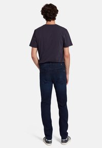 Vista posteriore di un uomo che indossa una t-shirt grigio scuro e jeans blu scuro. I jeans presentano una texture liscia e un design classico a cinque tasche. Scarpe casual completano il look.