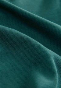 Žalsvai mėlynas audinys su smulkia, tekstūruota paviršiaus struktūra ir matomais briauneliais, išdėstytas minkštais klostėmis, atskleidžiantis lygų ir lengvą pobūdį.