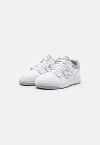 Para białych i szarych tenisówek New Balance z sznurówkami, perforowanym noskiem i gumową podeszwą na białym tle.