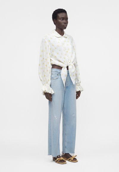 Γυναίκα με λευκό μπλουζάκι με κίτρινα σχέδια ήλιου, δεμένο μπροστά, με φουσκωτά μανίκια, ανοιζούμενα τζιν ανοιχτού μπλε και μπεζ σανδάλια με λουράκια-φιόγκους.