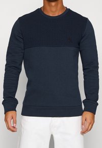 Sudadera azul marino con panel superior texturizado, cuello redondo y mangas largas. Presenta un pequeño logo oscuro en el frente. Material suave.