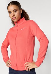 Tânără cu păr creț, îmbrăcată într-o jachetă corai cu fermoar, având logo-ul Champion, pozând cu mâinile pe șolduri, pe un fundal simplu.