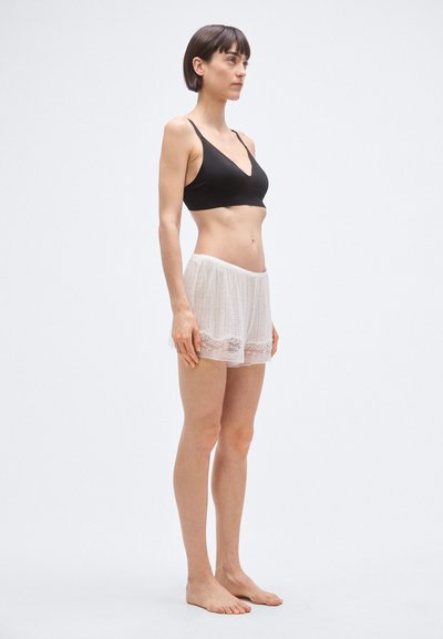 Črni bralette z tankimi naramnicami, kombiniran s prozornimi belimi kratkimi hlačami z čipkastim robom, ki jih nosi oseba, ki stoji v profilu.