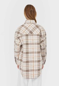 Chemise veste en flanelle avec un motif écossais beige et marron, col dans le cou, manches longues et ourlet arrondi. Elle présente une texture en laine propre et douce.