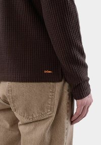 Brun ribbad tröja med långa ärmar, med en liten orange logodetalj i fållen; bärs tillsammans med beige, något löst sittande byxor.