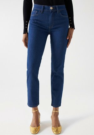 Femme portant un jean bleu taille haute associé à des talons dorés avec bride à la cheville et un top noir à manches longues avec détails de boutons.