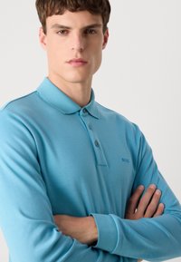Svetlo modra polo majica z dolgimi rokavi, ki ima dvonaborski izrez, rebrast ovratnik in diskretnim logotipom na prsih. Gladka tekstura, oprijet kroj.
