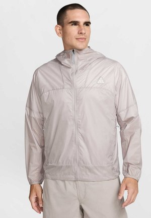 Giacca leggera con cappuccio, con zip frontale, di colore grigio chiaro. Realizzata in materiale liscio e lucido, con polsini elasticizzati e un logo triangolare sul petto.