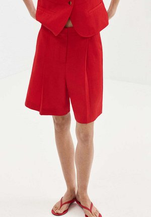 Persona que lleva pantalones cortos rojos hasta la rodilla, blazer rojo a juego y chanclas rojas, de pie sobre un fondo blanco.