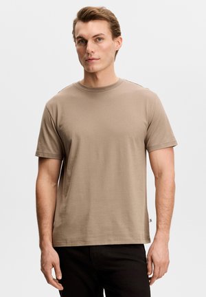 Junger Mann steht, trägt ein schlichtes beige kurzärmeliges T-Shirt und schwarze Hose, vor einem hellgrauen Hintergrund.