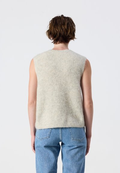 Personne aux cheveux bruns courts portant un pull en tricot beige sans manches et un jean bleu, vue de dos sur un fond uni.