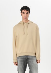 Sweatshirt bege com capuz em tecido macio, apresentando um cordão, punhos e bainha canelados. Combinado com uns jeans cinza claro.