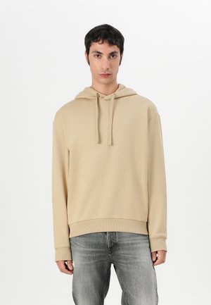 Beige Kapuzenpullover aus weichem Stoff, mit Kordelzug, gerippten Bündchen und Saum. Kombiniert mit hellgrauen Jeans.