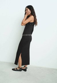 Robe noire sans manches avec un corsage ajusté, une fente sur le côté et un accent argenté à la taille. Portée avec des chaussures plates noires dotées de brides à la cheville et de matériel.