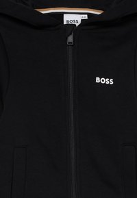 Casaco com fecho éclair preto, feito de tecido macio, com um fecho na frente, dois bolsos laterais e um logótipo branco "BOSS" no lado esquerdo do peito.