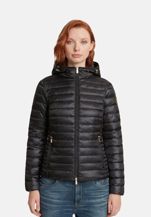 Frau mit roten Haaren, die eine schwarze, gesteppte Kapuzenjacke und blaue Jeans trägt, steht vor weißem Hintergrund.