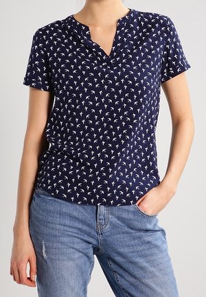 Femme portant une chemise marine à manches courtes avec des motifs d'oiseaux blancs et un jean bleu clair, une main dans la poche, sur un fond uni.