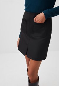 Gonna mini nera con uno spacco frontale e due tasche laterali, realizzata in un tessuto liscio e con un taglio sartoriale. Indossata con un top a maniche lunghe color teal.