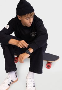 Sweatshirt preto de gola redonda com logótipo, calças de ganga escuras, ténis brancos com riscas pretas e um gorro preto. Skateboard com rodas vermelhas.