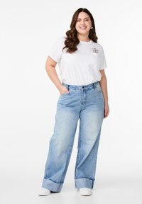Bílý tričko s malým kapsičkovým designem, spárované se světle modrými širokými džíny s ohrnutým lemem. Model má na sobě bílé tenisky.