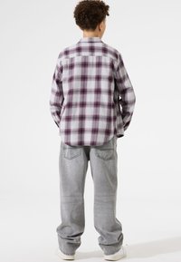 Persoon met krullend haar staat met de rug naar de camera, gekleed in een wit en paars geruit shirt, grijze jeans en witte sneakers op een effen achtergrond.