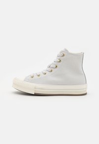 Høyde sneakers laget av lys grå kanvas, med gulløyer, en hvit gummitåhette og en teksturert hvit såle med brune detaljer.