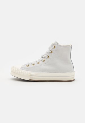 Høyde sneakers laget av lys grå kanvas, med gulløyer, en hvit gummitåhette og en teksturert hvit såle med brune detaljer.