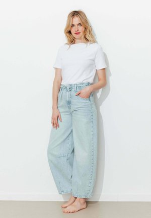 Femme blonde debout pieds nus contre un mur blanc, portant un t-shirt blanc uni et un jean bleu clair à jambes larges avec une ceinture à nouer.