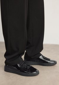 Baskets en cuir verni noir avec un bout arrondi, un design à lacets et une semelle en caoutchouc, placées à côté d'un pantalon noir ample.