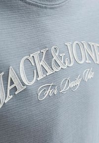 Ανοιχτό μπλε ύφασμα με ανάγλυφο λευκό κείμενο "JACK & JONES" και καλλιγραφικό "For Daily Use" τυπωμένο στο μπροστινό μέρος.
