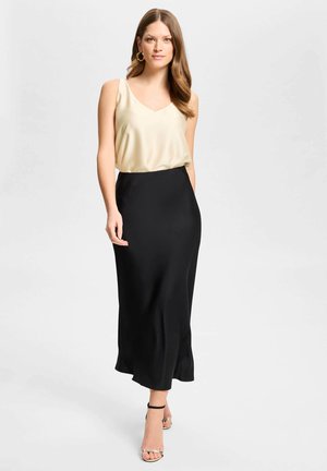 Frau mit langen braunen Haaren, die ein ärmelloses goldenes Oberteil, einen schwarzen Midirock und goldene High Heels trägt, stehend auf weißem Hintergrund.