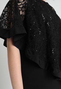 Schwarze Spitzenbluse mit floralen Mustern, Pailletten und Rüschensäumen, getragen von einer Person mit einem sichtbaren, nackten Arm vor einem grauen Hintergrund.