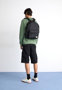 Sort rygsæk med en glat overflade, der har en frontlomme og et logo-patch, båret af en person i en grøn sweatshirt og sorte shorts.