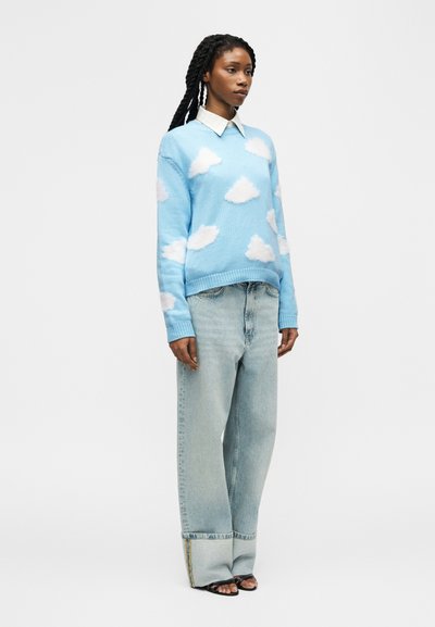 Femme aux cheveux tressés portant un pull bleu avec des nuages blancs, une chemise blanche à col, un jean clair à jambes larges, et des sandales noires, debout de profil.