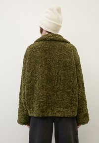 Olivengrøn, fuzzy yderjakke med bred krave og lange ærmer, parret med en cremefarvet hue; tekstureret stof med et blødt udseende.