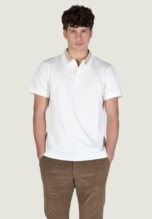 Biała koszulka polo z krótkim rękawem, beżowym kołnierzem i troje guzikami, w połączeniu z brązowymi spodniami sztruksowymi, ręce w kieszeniach.