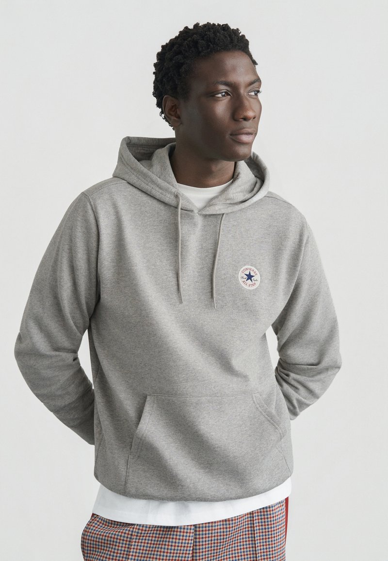 Grå hættetrøje lavet af et blødt materiale, med en frontlomme og en cirkulær logopatch. Lag-på-lag over en hvid skjorte med en struktureret kant.