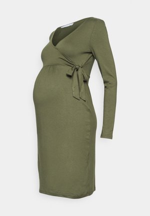 Długa sukienka ciążowa w kolorze oliwkowej zieleni, z długimi rękawami, z przodu z zakładką i wiązaniem z boku, o długości do kolan, na jednolitym jasnym tle.