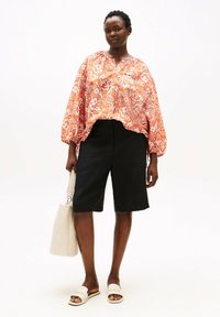 Flydende orange og hvid blomsterbluse med lange ærmer, kombineret med sorte shorts til knæet. Et let håndtaske og sandaler fuldender looket.