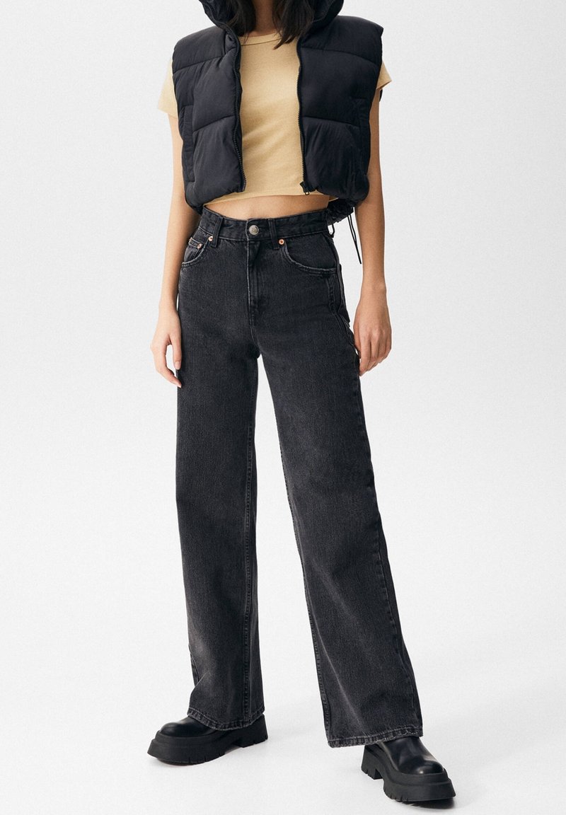 Žena v černé péřové vestě přes béžový crop top, v černých vysokých širokých džínách a černých platformových botách, stojící na bílém pozadí.