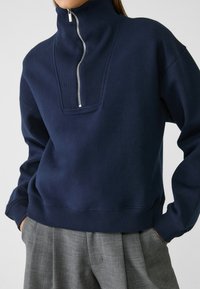 Marineblauwe sweatshirt met een hoge kraag, halve ritssluiting en geribbelde accenten. Heeft een losse pasvorm en zachte textuur, gecombineerd met grijze, op maat gemaakte broek.