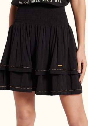 Jupe noire à étages avec tissu texturé et petits détails de perles dorées le long de l'ourlet, portée par une personne la main reposant le long du corps.