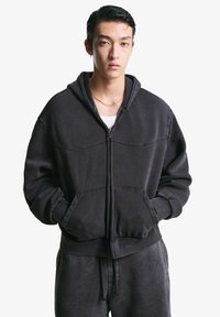 Junger Mann in einem schwarzen Zip-Hoodie und passenden Hosen, mit Händen in den Taschen, einer goldenen Kette um den Hals vor einem schlichten weißen Hintergrund.