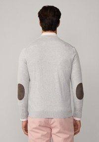 Grauer Pullover mit braunen Ellbogen-Patches, gestrickter Textur und gerippten Bündchen, getragen über einem hellrosa Hemd und kombiniert mit hellrosa Hosen.