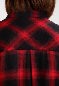 Chemise à carreaux rouge et noire avec un col, un tissu texturé et un pli dans le dos.