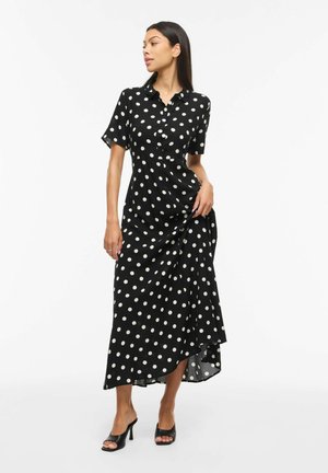 Sieviete tērpusies melnā, īsroku, ar pogām, potītēm garā kleitā ar baltām polka punktu zīmēm un melniem atvērtu purngalu augstpapēžu kurpēm, stāv uz balta fona.