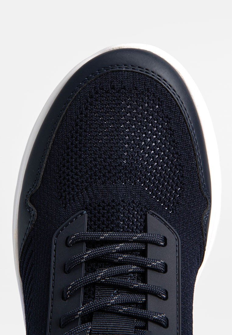 Zbliżenie czarnej dzianinowej tenisówki z okrągłym czubkiem, czarnymi sznurówkami i białą gumową podeszwą na jednolitym tle.