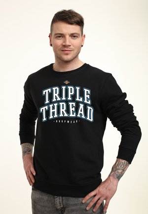 Mand med kort hår og tatoveringer iført en sort "TRIPLE THREAD" sweatshirt, stående med hænderne på hofterne mod en ensartet lys baggrund.